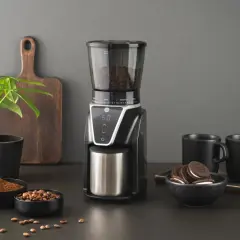 En modern kaffekvarn med digital display, fylld med bönor, bredvid skålar med malet kaffe och hela bönor. Två svarta muggar och en liten krukväxt fulländar scenen.