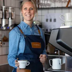 En leende barista håller en kaffekopp. Kanske en påminnelse om hur mycket kaffe dricker svenskar?