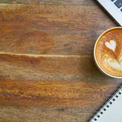 En kopp kaffe med lattekonst, en laptop och en anteckningsbok på ett träbord. Perfekt för att få lite koffein och inspiration.