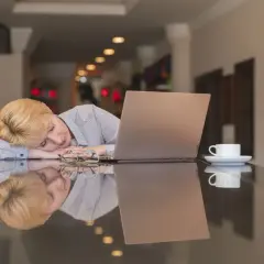 Kvinnan med blont hår sover vid ett bord med en laptop och en kaffekopp. Hon verkar trött, trots att hon inte blir pigg av kaffe.