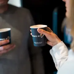 Två personer håller kaffemuggar. En kvinna håller en mugg med texten "Beans in Up". Kanske är det här svaret på vilket land dricker mest kaffe.