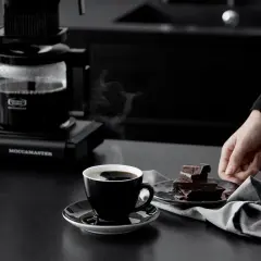 En hand sträcker sig efter chokladbitar bredvid en kopp ångande kaffe från en Moccamaster. Kanske är det dåligt kaffe, men chokladen ser god ut.