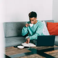 Ung man sitter i soffan med en kopp, kanske en mate drink, och läser en bok. En laptop står på bordet.