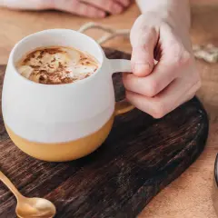 En hand håller en mugg med en krämig pumpa latte, toppad med kanel. En guldig sked ligger bredvid.