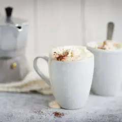 Två muggar med krämig mocca kaffe, toppad med vispgrädde och kakao. En mokabryggare står i bakgrunden, redo för ett nytt mocca kaffe recept.