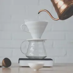 Vatten hälls från en kopparkanna över ett filter i en vit V60-bryggare, som står på en glaskaraff. En kaffekvarn och en skopa ligger bredvid. Detta visar hur brygger man kaffe.