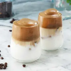 Två glas med krämigt dalgona kaffe, med en fluffig kaffetopping över mjölk och is. Kaffebönor sprider sig på marmorbordet.