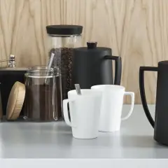 Kaffebönor och malet kaffe i glasburkar, en kaffekvarn, två vita muggar och två svarta presskannor. Perfekt för att utforska olika presskaffe sorter.