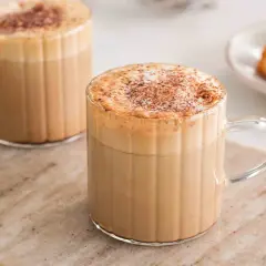 Två glas med krämigt skummande cafe americano, toppade med kanel. Bredvid står en liten tallrik med gyllene kokoskakor.