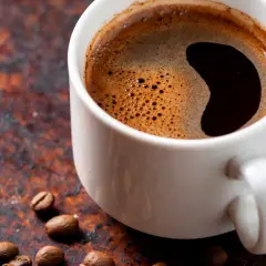 En vit kopp med starkt kaffe och skummig yta, omgiven av kaffebönor på en rustik bakgrund.