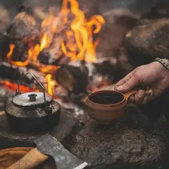 Vid en sprakande eld, med en kaffepanna och en träkopp, kan man brygga kokkaffe. En yxa och handskar ligger bredvid.