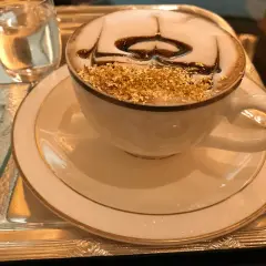 En lyxig kaffekopp med guldfoliering och chokladdrizzle, kanske det dyraste kaffet någonsin. Serveras på ett silverfat med ett glas vatten.