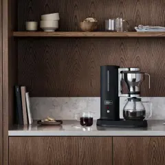 En stilren kaffebryggare i svart och krom, bredvid en glaskaraff med kaffe. På hyllan ovanför finns skålar och glas. En av flera olika kaffebryggare som passar in i ett modernt kök.