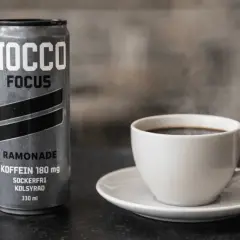En burk NOCCO Focus Ramonade med 180 mg koffein bredvid en ångande kopp kaffe. Perfekt för att kickstarta dagen med extra espresso koffein.