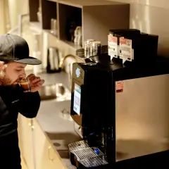 Man med keps njuter av en espresso i fikarummet på arbetsplatsen. En modern kaffemaskin står redo för nästa kopp.