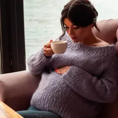 Pregnant woman in a purple sweater holding a cup of tea, resting her hand on her belly.