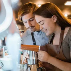 Två personer lär sig barista-yrket vid en espressomaskin. Hon håller en mjölkkanna, han tittar på.