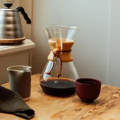 En Chemex med färdigbryggt dropp kaffe står på ett träbord. Bredvid finns en liten kanna och en kopp. En vattenkokare syns i bakgrunden.