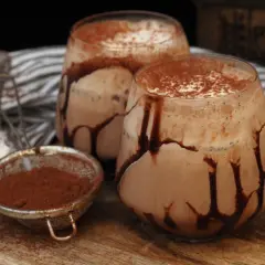 Två glas med krämig kaffe med choklad, garnerade med chokladsås och kakaopulver. En sil med kakaopulver står bredvid.