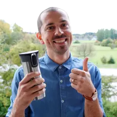 Glad man med tummen upp håller en termosmugg, redo för sitt fett kaffe utomhus.