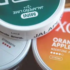 Tre dosor nikotinpåsar: en grön med jalapeño lime, en vit med texten "strength" och en orange med smak av apelsin och äpple. Frågan "hur många prillor i en dosa" kan besvaras genom att titta på dessa.