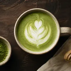 En kopp vackert dekorerad matcha latte med ett hjärtmönster, bredvid en skål med matchapulver och en bambuvisp.