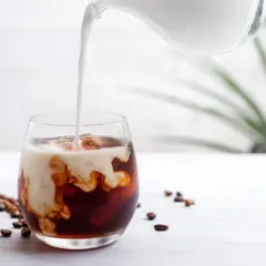 Mjölk hälls i ett glas med iskaffe på snabbkaffe. Kaffebönor ligger utspridda på bordet.