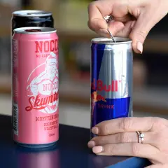 En hand öppnar en burk Red Bull, bredvid NOCCO-burkar. Koffein redbull för energi.