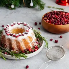 En vackert dekorerad sockerkaka med lingon och rosmarin, perfekt för julfika till många. En skål med lingon och en kopp te står redo.