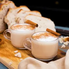 Två muggar krämig chai latte med kanelstänger och strödd kanel. Bröd och kryddor i bakgrunden. Perfekt för ett mysigt chai latte recept.
