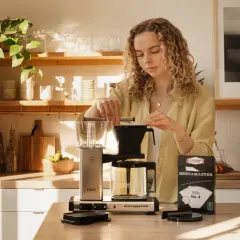 En kvinna förbereder kaffe med en Moccamaster. På bordet ligger en kartong med kaffefilter, där det står "100x No. 4". Det ger en ledtråd om hur många kaffemått per kopp som kan behövas.