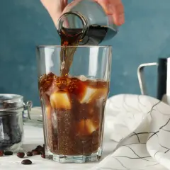 En hand häller mörkt kaffe över is i ett glas. Bredvid står en burk med kaffebönor. Kaffe smakar surt om det inte är perfekt bryggt.
