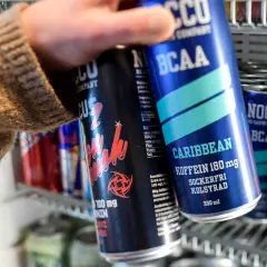 En hand tar en NOCCO Caribbean ur ett kylskåp fyllt med energidrycker. Frågan "hur många nocco per dag" dyker upp när man ser alla dessa.