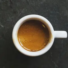 En vit kopp med en gyllene caffe macchiato med crema på en mörk, stenig yta.