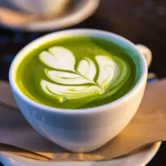 En vit kopp med grön matcha latte, dekorerad med ett vitt mönster som liknar ett löv. Matcha innehåll i sin finaste form.