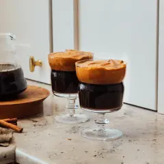 Två glas med mörkt, kallt kaffe toppat med fluffigt skum. En karaff med kaffe och kanelstänger står bredvid.