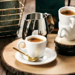 Enkel espresso i vit kopp med guldfärgad sked. En mjölkkanna och en stapel koppar står bredvid.