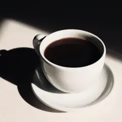En vit kaffekopp med mörkt kaffe, belyst av solljus som skapar dramatiska skuggor. Hur påverkar kaffe blodtrycket?