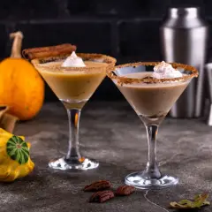 Två martiniglas med krämig pumpkin spice latte, garnerade med vispgrädde och kanelstång. Pumpan och höstlöv skapar en mysig stämning.