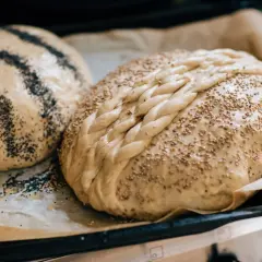 Två nybakade bröd på bakplåtspapper, ett toppat med vallmofrön och ett med sesamfrön och flätat mönster. Perfekta fika idéer!