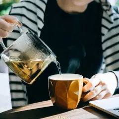 En person häller upp ångande, nyttigt te från en glaskanna i en orange mugg. En laptop syns bredvid.