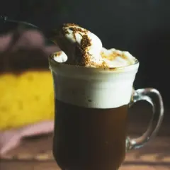 En sked med vispad grädde och kanel toppar en varm kaffe Karlsson utan Baileys. I bakgrunden syns en gul kaka.