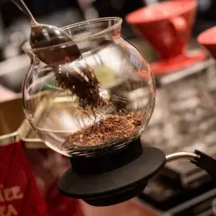 En sked lägger teblad i en glaskaraff, redo för att brygga te utan koffein. Röda kaffefilter står i bakgrunden.