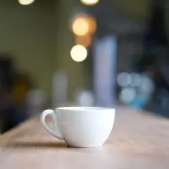 En vit, tom mått kaffekopp står på ett träbord. Bakgrunden är suddig med ljus.