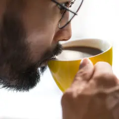 Man med glasögon dricker kaffe ur en gul mugg. Koffein ger energi, motverkar adenosin.