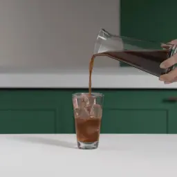 En hand häller kallt kaffe med isbitar i ett glas.
