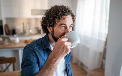 Man med lockigt hår dricker kaffe, blicken fundersam. Kanske en stund av kaffe ångest innan dagen börjar.
