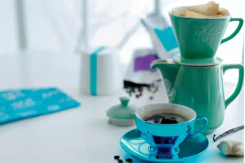En turkos kaffekopp med fat står framför en grön kaffekanna med filterhållare. Kaffebönor och en vaniljstång ligger bredvid. En fin morgon med gott kaffe.