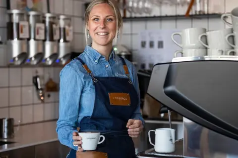 En leende barista håller en kaffekopp. Kanske en påminnelse om hur mycket kaffe dricker svenskar?