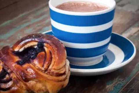 En blårandig kopp med kaffe och en kanelbulle med blåbär. Perfekt för en fika till jobbet.